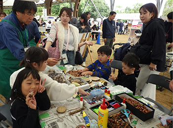 イベントブース木工工作のにぎわい立った大人男女3人。座った男女幼児４人