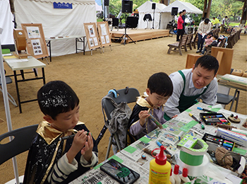 イベントブース木工工作で細かい作業する男幼児2人と緑のエプロン成人男性1人