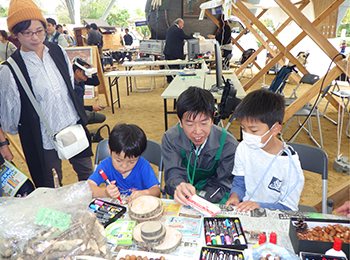 イベントブース木工工作のにぎわい立った成人女1人。座った男幼児2人の間に成人男性1人