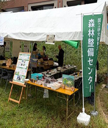 イベントテント外観。事業説明パネルと作業等のための机上に資材が並ぶ。整備センターの緑地に白文字の旗。