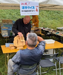 イベントテントの外縁部で木偏の漢字クイズ。挑戦する初老男性右手で頭を抱える。グレーのワークシャツ