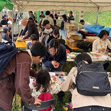 イベントテントの内のにぎわい。子供一人に夫婦手前から熱心に手元見る母親らしき黒服女性と白セーター成人女性他奥のテーブルも参加多数