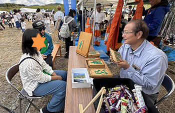 横から見た木の漢字あてクイズ。左に挑戦者小学校高学年女子茶色ストライプシャツ青パンツ。出題者50代男性笑顔。遠景に青空