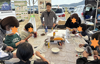 イベントテント内クリスマスツリー作業机縁に座る小学校低学年児童男女5人。左手に保護者成人女性グレーの服左と黒パーカー右手。中央に立つグレー作業着職員