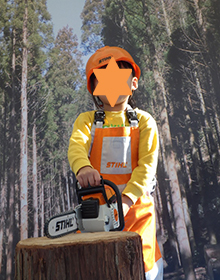 オレンジのヘルメットと防護服で林業従事者に扮した5歳くらいの児童、手には模型の電動ノコギリ