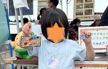 イベントテント入り口で作成した木製ストラップを嬉しそうに掲げる3歳くらいの女児全身。水色シャツに柄黄色パンツ白い靴。