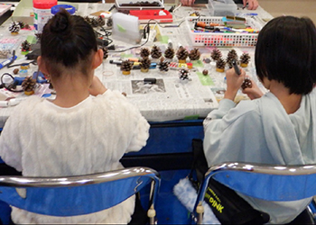 工作テーブルで作業する小学校低学年女児2名松ぼっくり多数
