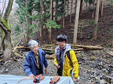 屋外の森林整備現場で青い服の林業関係者と打ち合わせる黄色いジャケット寺口職員。背後に造林木。写真