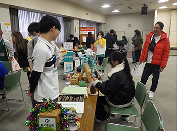 会場で木へんのつく漢字クイズを真横から。左手白シャツ職員。中央挑戦する焦げ茶ジャケット女性。背後に赤ダウン男性