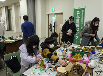 にぎわう松ぼっくりクリスマスツリー作業机。小3くらい男女児童、センター旗付近成人女性女性2人