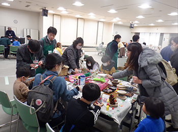 にぎわう松ぼっくりクリスマスツリー作業机別角度。手前低学年2名と父親、右にグレーダウン着た母親資材を探す