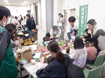 にぎわう松ぼっくりクリスマスツリー作業机別角度。乳幼児抱いて工作の母親手前。右に小学校低学年女児2名と白ダウン母親他職員補助2名周囲